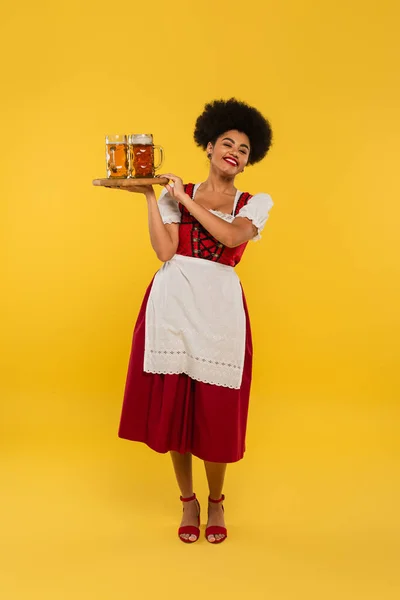 Pleased african american bavarian waitress holding wooden tray with beer mugs on yellow, banner — Stock Photo