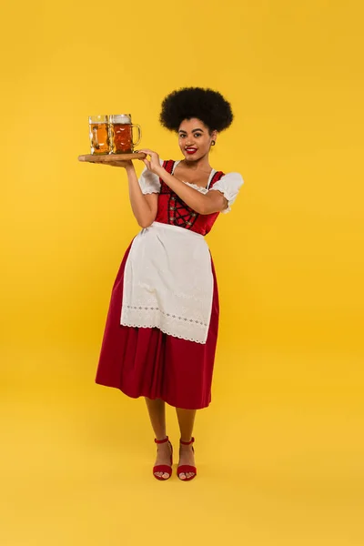 Smiling african american oktoberfest waitress holding wooden tray with beer mugs on yellow, banner — Stock Photo