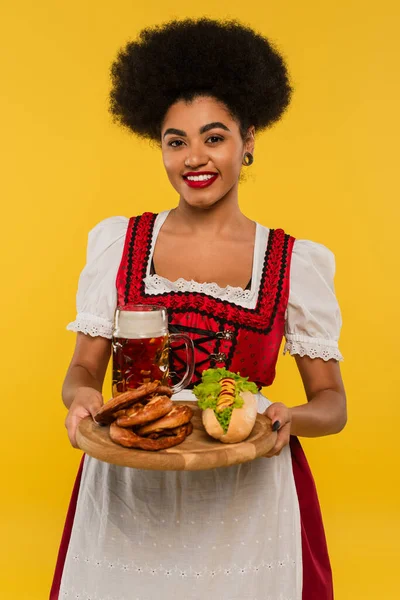 Alegre africano americano oktoberfest camarera sosteniendo bandeja de madera con pretzels y hot dog en amarillo - foto de stock