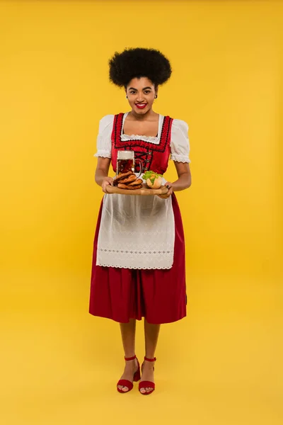 African american oktoberfest waitress holding wooden tray with beer and delicious treat on yellow — Stock Photo