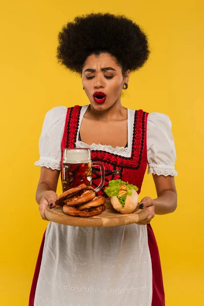 Amazed african american bavarian waitress with beer and delicious treat on wooden tray on yellow — Stock Photo