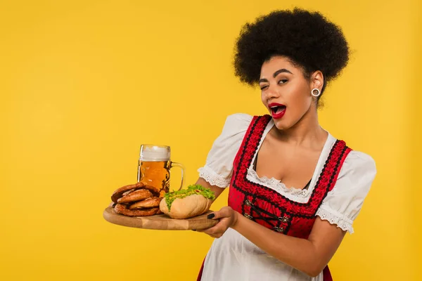 African american oktoberfest waitress with beer and snacks on wooden tray winking on yellow, banner — Stock Photo