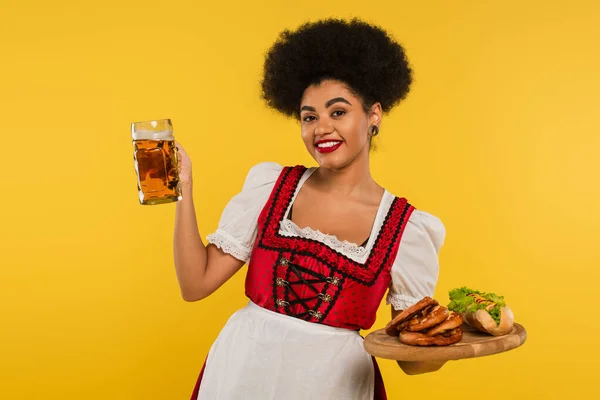 Feliz africano americano oktoberfest camarera con cerveza y comida en bandeja de madera en amarillo, bandera - foto de stock