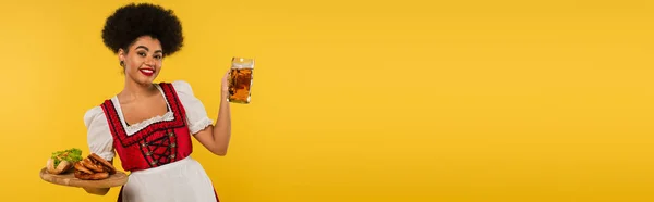 Serveuse bavaroise afro-américaine souriante tenant de la bière et de la nourriture sur un plateau en bois jaune, bannière — Photo de stock