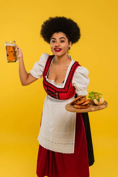Молода афроамериканська баварська офіціантка в рендлі, що подає пиво та їжу на дерев'яному лотку на жовтому — стокове фото