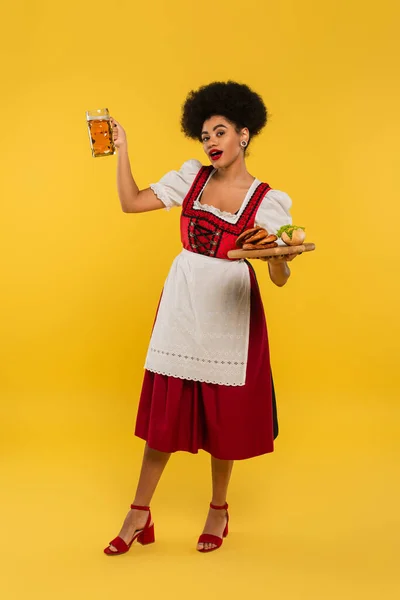 African american bavarian waitress in dirndl posing with beer and food on wooden tray on yellow — Stock Photo