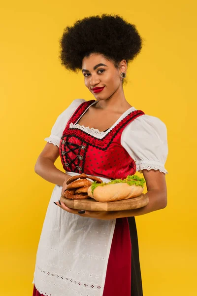 Charming african american oktoberfest waitress with pretzels and hot dog on wooden tray on yellow — Stock Photo