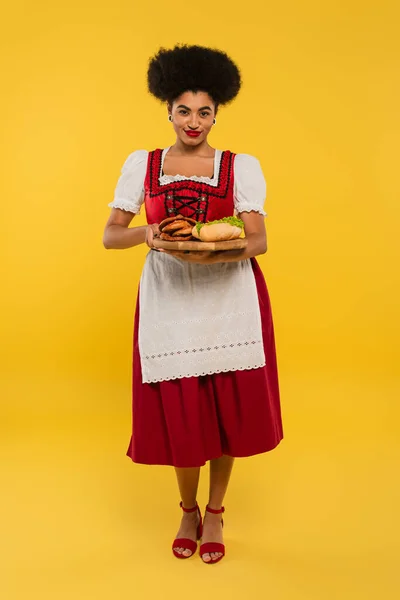 Pleased african american bavarian waitress serving pretzels and hot dog on wooden tray on yellow — Stock Photo