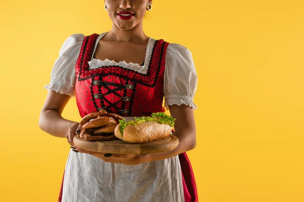 Cropped african american bavarian waitress with pretzels and hot dog on wooden tray on yellow — Stock Photo
