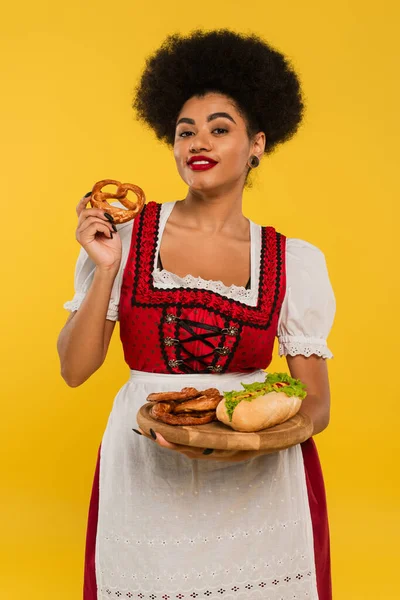 Joyful african american oktoberfest waitress holding pretzels and hot dog on wooden tray on yellow — Stock Photo
