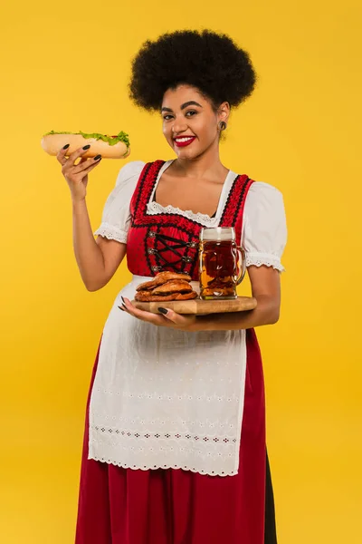 Cheerful african american bavarian waitress with beer, pretzels and hot dog on wooden tray on yellow — Stock Photo