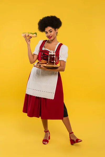 Joyful african american oktoberfest waitress with beer and delicious treat on wooden tray on yellow — Stock Photo