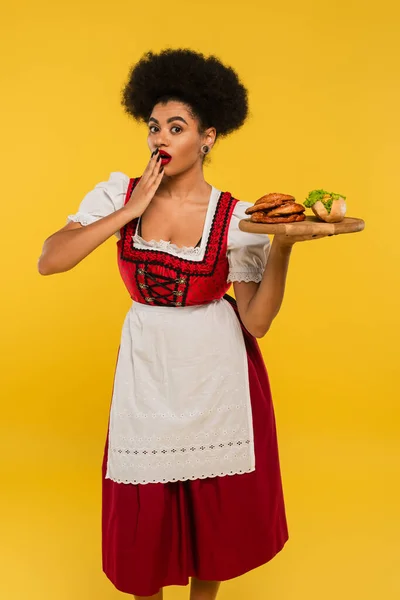 Шокированная африканская американская баварская официантка держит деревянный поднос с крендельками и хот-догом на желтом — стоковое фото
