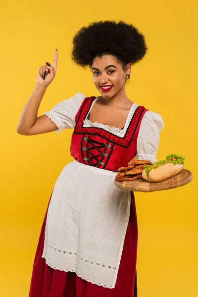 Joyful african american oktoberfest waitress with snacks on wooden tray showing idea sign on yellow — Stock Photo