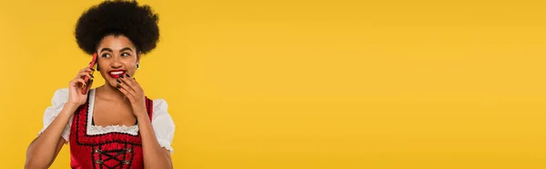 Felice cameriera bavarese africana americana in abbigliamento autentico parlando su smartphone su giallo, banner — Foto stock