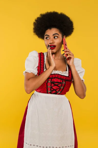 Giovane afro americano oktoberfest cameriera in Dirndl costume pettegolezzi su smartphone su giallo — Foto stock