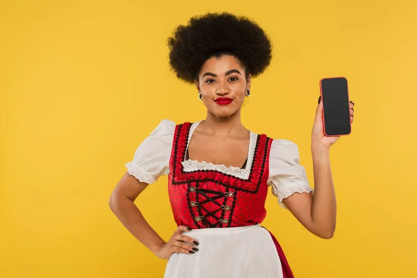 Serveuse afro-américaine oktoberfest en dirndl montrant smartphone avec écran blanc sur jaune — Photo de stock