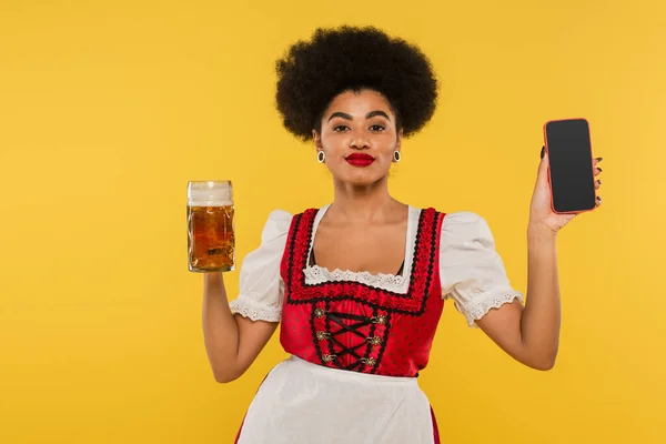 Camarera bavariana afroamericana con taza de cerveza y teléfono inteligente con pantalla en blanco en amarillo - foto de stock
