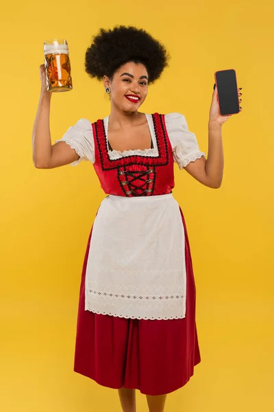 Felice cameriera bavarese africana americana con tazza di birra e telefono cellulare con schermo bianco su giallo — Foto stock