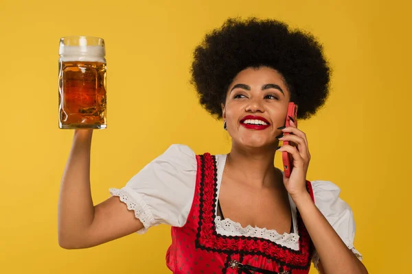 Aufgeregte afrikanisch-amerikanische Oktoberfest-Kellnerin mit Bierkrug redet auf Smartphone auf gelb — Stockfoto