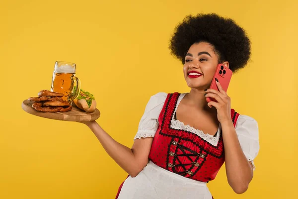 Camarera bavariana afroamericana con cerveza y aperitivos en bandeja hablando en teléfono inteligente en amarillo - foto de stock