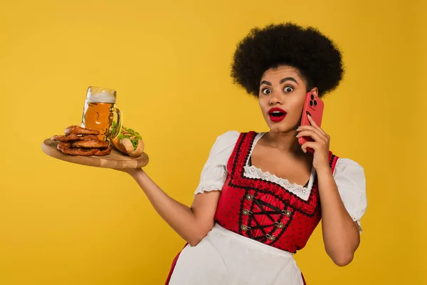 Surprise serveuse bavaroise afro-américaine avec bière et snacks parlant sur smartphone sur jaune — Photo de stock