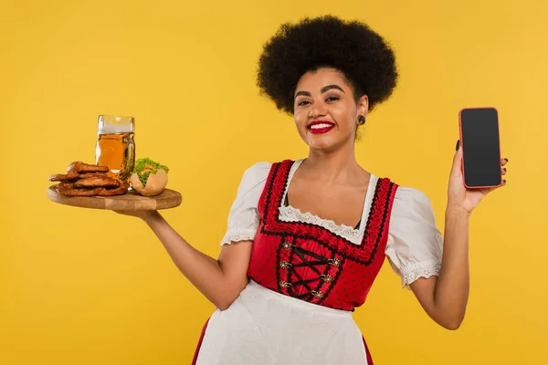 African american bavarian waitress with beer and snacks showing blank screen of smartphone on yellow — Stock Photo