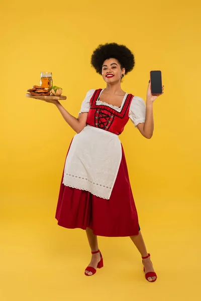African american bavarian waitress with beer and food showing blank screen of smartphone on yellow — Stock Photo