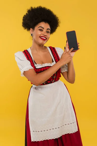 Happy african american bavarian waitress in dirndl pointing at blank screen of smartphone on yellow — Stock Photo