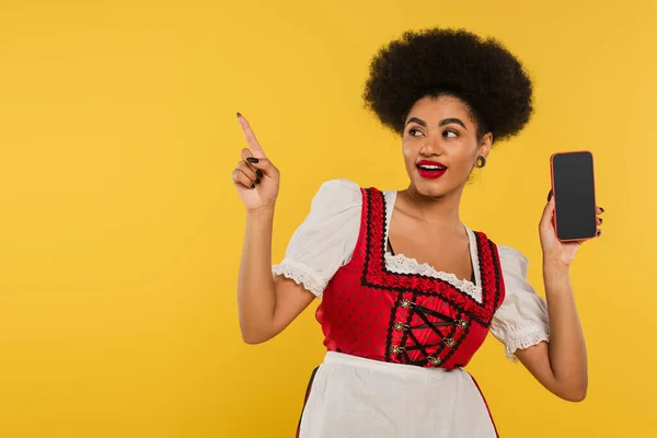 African american bavarian waitress with smartphone pointing with finger, showing direction on yellow — Stock Photo