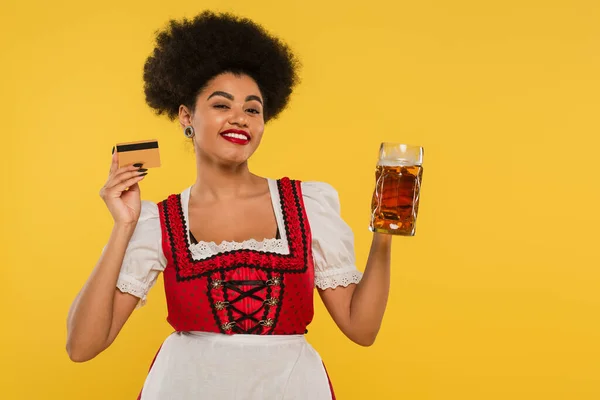 Gioiosa cameriera bavarese africana americana in costume Dirndl con tazza di birra e carta di credito su giallo — Foto stock