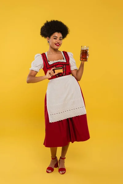 Feliz Africano americano bavarian garçonete em dirndl vestido com caneca de cerveja e cartão de crédito no amarelo — Fotografia de Stock