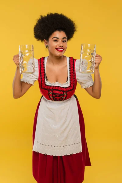 Heureuse serveuse afro-américaine oktoberfest en robe dirndl tenant des tasses de bière vides sur jaune — Photo de stock