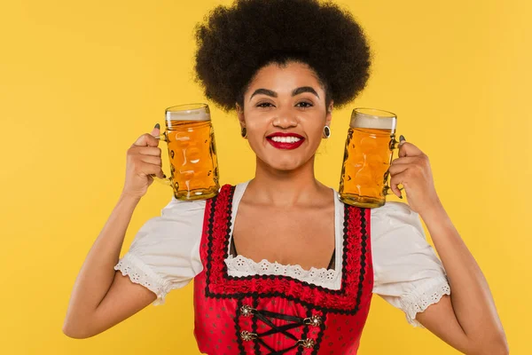 Smiling african american oktoberfest waitress in dirndl dress holding mugs with craft beer on yellow — Stock Photo