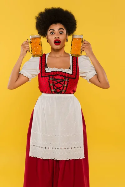 Garçonete americana africana maravilhada em traje tradicional bávaro segurando canecas de cerveja no amarelo — Fotografia de Stock