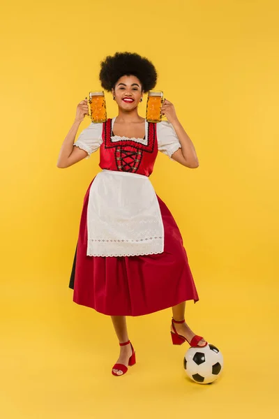 Cheerful african american waitress in dirndl standing with beer mugs near soccer ball on yellow — Stock Photo