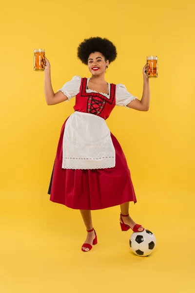 Fröhliche afrikanisch-amerikanische Kellnerin im Dirndl mit Bierkrügen in der Nähe des Fußballs auf gelb — Stockfoto