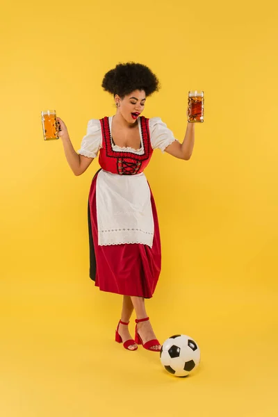 Радісна афроамериканська баварська офіціантка в дирндлі тримає пивні чашки біля футбольного м'яча на жовтому — стокове фото