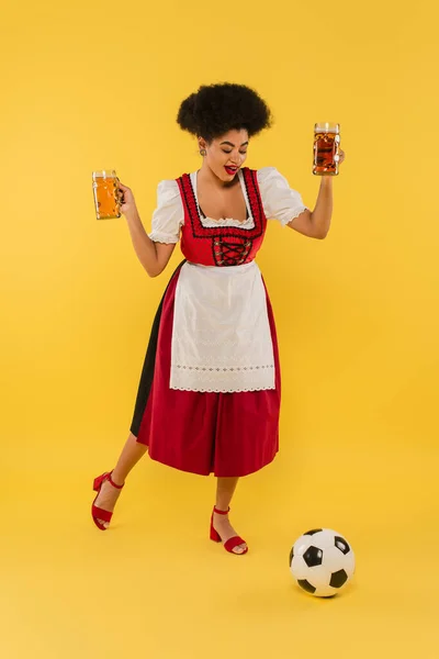 Joyous african american oktoberfest waitress holding beer mugs and playing football on yellow — Stock Photo