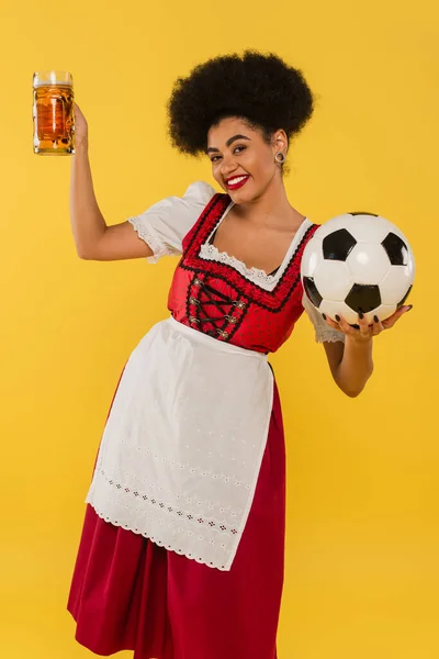 Unbekümmerte afrikanisch-amerikanische Oktoberfestkellnerin im Dirndl mit Bierkrug und Fußball auf gelb — Stockfoto