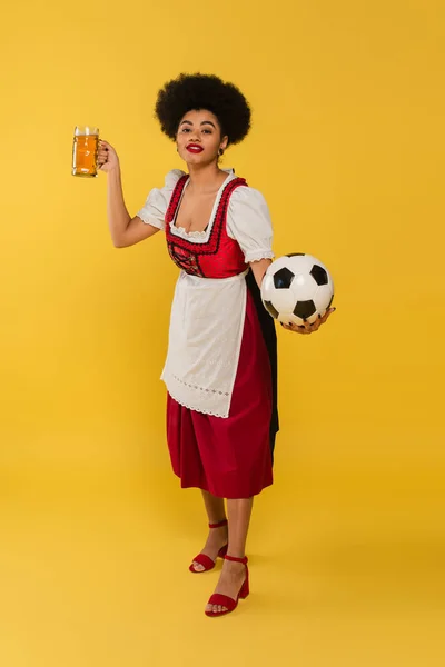 Erfreut afrikanisch-amerikanische Kellnerin in bayerischer Tracht mit Bierkrug und Fußball auf gelb — Stockfoto