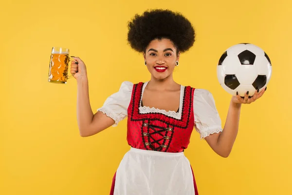 Cheerful african american bavarian waitress in dirndl holding beer mug and soccer ball on yellow — Stock Photo