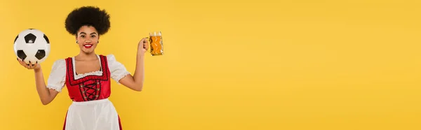 Happy african american bavarian waitress in dirndl with beer mug and soccer ball on yellow, banner — Stock Photo