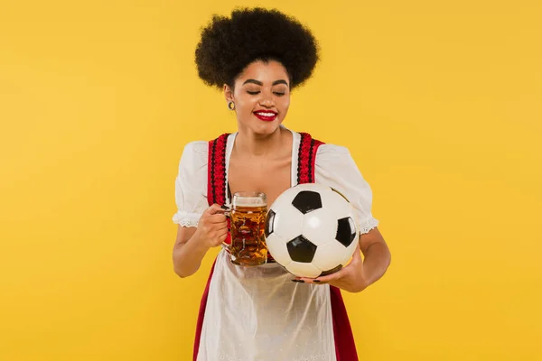 Joyful african american oktoberfest waitress in dirndl clinking beer mug with soccer ball on yellow — Stock Photo