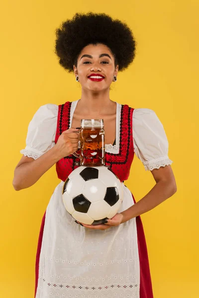 Happy african american bavarian waitress with beer mug and soccer ball smiling on yellow — Stock Photo