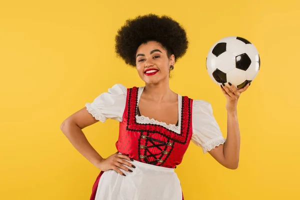Encantadora camarera bavariana afroamericana posando con pelota de fútbol y mano en la cadera en amarillo - foto de stock