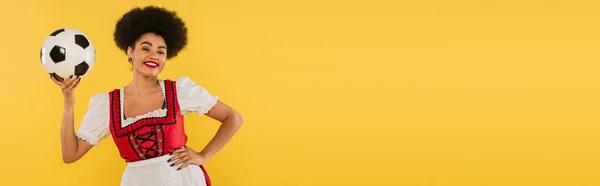 Happy african american bavarian waitress posing with soccer ball and hand on hip on yellow, banner — Stock Photo