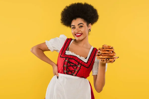 African american oktoberfest waitress in traditional costume holding pretzels and smiling on yellow — Stock Photo