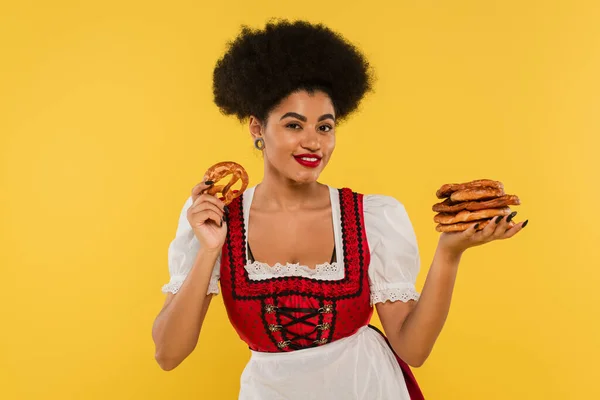 Joyful african american oktoberfest waitress in authentic costume holding pretzels on yellow — Stock Photo