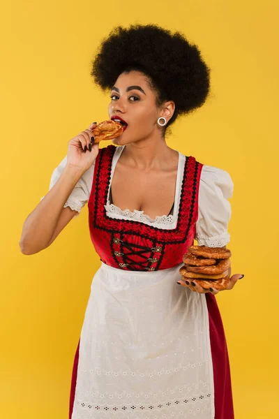 Encantadora garçonete afro-americana em traje oktoberfest comendo delicioso pretzel em amarelo — Fotografia de Stock